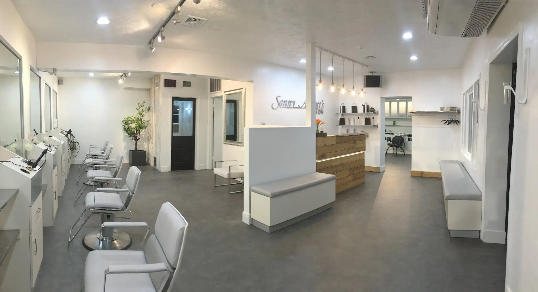 A row of white and chrome styling chairs, a pine reception desk and Edison pendant lamps in a bright white room.