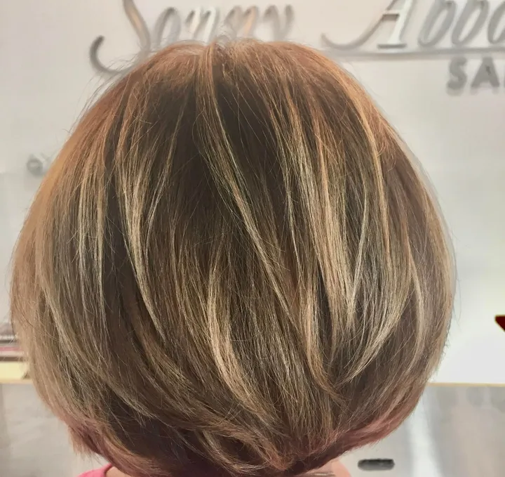 Rounded bob with caramel highlights, seen from behind, in front of the salon’s brushed metal lettering.