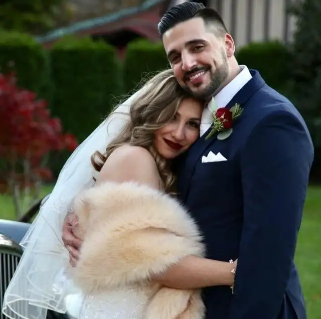 Bride with soft blonde waves and a veil, held by her groom in a navy suit with a skin fade and beard.